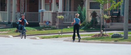 A paperboy rides his bike and throws a newspaper, whilst a female jogger runs past