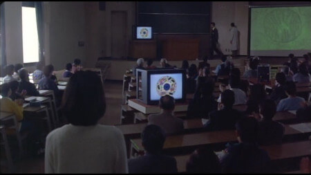 A packed lecture theatre, with Kiyomi looking on, as images of Mitochondria are shown